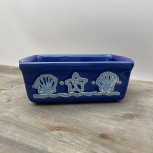 May include: A rectangular, cobalt blue ceramic dish with a coastal theme. The dish features light blue seashell and starfish designs above a wavy line. The design is centered on the front of the dish.