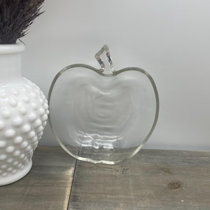 May include: A clear glass apple-shaped dish with a stem detail. The dish is sitting on a wooden surface next to a white vase with a textured surface. The background is a plain white wall.