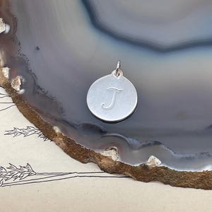 May include: A round, silver-colored pendant with the letter "J" engraved on its surface. The pendant is suspended from a small loop at the top. The background is a gray and brown stone.