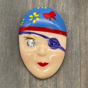 May include: A vintage pirate-themed face brooch. The brooch features a blue hat with yellow and red accents, a red stripe, and a blue eye patch. The face has a sparkling rhinestone eye and red lips. The brooch is made of a glossy material.