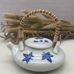 May include: A small white ceramic teapot with a blue and white maple leaf design. The teapot has a bamboo handle and a lid with the text "豊穣" in black.