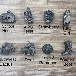 May include: Ten pewter charms with different designs, including a schoolhouse, a story teller, a strawberry, a tulip girl, a southwest cactus, a deer, a love and romance symbol, and a western boot.