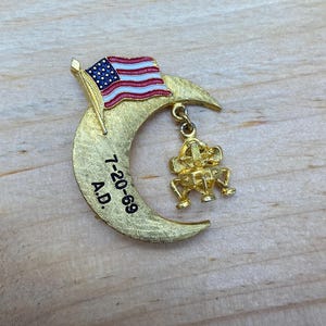 Op de afbeelding: Een gouden halve maan broche met een Amerikaanse vlag en een klein model van de maanlander die eraan hangt. De maan heeft de datum "7-20-69 A.D." erop gegraveerd.