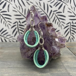 Pode incluir: Um par de brincos turquesa em forma de gota com detalhes em metal prateado. Os brincos são exibidos em um cristal roxo contra um fundo padronizado. Os brincos têm uma aparência de couro trançado.