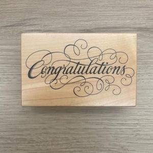 May include: A rectangular wooden rubber stamp featuring the word Congratulations in a black cursive script font with decorative flourishes and swirls surrounding the text, set against a light brown natural wood grain background.