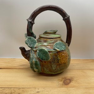 May include: A ceramic teapot with a brown handle and a green lid. The body of the teapot is decorated with green leaves and a cluster of yellow-brown fruit. The teapot is set on a wooden surface.