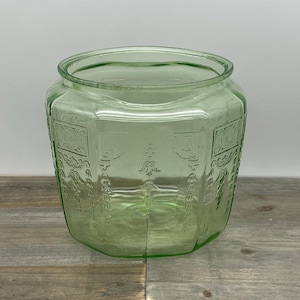 May include: A light green, octagonal glass jar with embossed decorative panels. The jar has a wide opening and sits on a wooden surface. The jar's design features repeating patterns and is likely vintage.