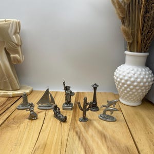 May include: A collection of ten pewter figurines, including a lighthouse, a sailboat, the Statue of Liberty, a cactus, the Space Needle, a sleigh, and two dolphins. The figurines are arranged on a wooden surface.