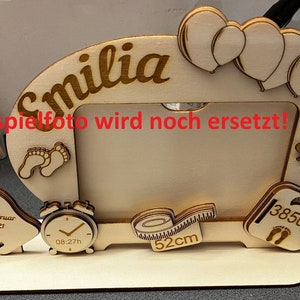 Può includere: Una cornice per foto in legno con un ritaglio di un'impronta di piede di bambino, una bottiglia, un orologio e un metro a nastro. La cornice è decorata con palloncini e il nome "Emilia" è scritto in alto. Il testo "Beispielfoto wird noch ersetzt!" è scritto sulla cornice. La cornice è su una superficie bianca.