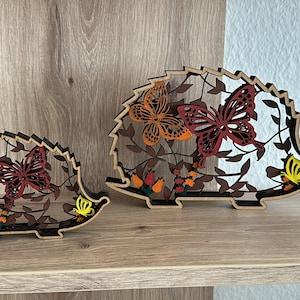 May include: Two wooden hedgehog figurines with butterfly and floral cutouts. The hedgehogs are brown with orange, red, and yellow accents. They are displayed on a wooden shelf against a light-colored wall.