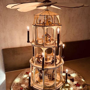 Op de afbeelding: Een houten kerstpiramide met drie niveaus, met miniatuurhuisjes, bomen en figuren. Het heeft kaarshouders en een draaiende top. De scène wordt verlicht door kaarsen en omgeven door feestelijke verlichting en decoraties.