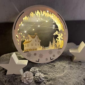 Peut inclure: Une scène de village de Noël en bois blanc avec une maison, des arbres et des figurines. La scène est éclairée par des lumières blanches et est entourée de deux ornements en forme d'étoile blanche. La scène est sur une surface grise et duveteuse.