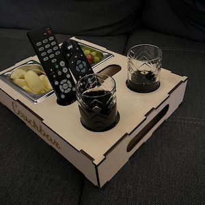 Könnte beinhalten: Ein Holztablett mit dem eingravierten Text "Couchbar" an der Seite. Das Tablett hat Fächer für Snacks, Getränke und Fernbedienungen. Es enthält zwei Gläser, eine Schüssel mit Chips und zwei Fernbedienungen.
