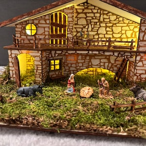 Può includere: Un presepe in miniatura con un edificio dalla texture in pietra, illuminato dall'interno. La scena comprende statuine di Maria, Giuseppe e Gesù bambino in una mangiatoia, insieme ad animali e una staccionata in legno. Il tema è religioso.