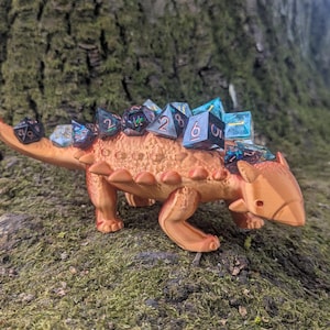 May include: A 3D printed orange ankylosaurus dinosaur figurine with a row of black and blue iridescent dice on its back. The dinosaur is standing on a bed of green moss.