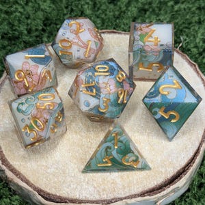 May include: A set of seven polyhedral dice for tabletop games. The dice have a floral design with pink and blue accents, and gold numbering. The dice are displayed on a wooden slice.
