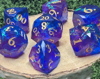 May include: A set of seven polyhedral dice featuring a translucent blue and purple gradient with embedded iridescent foil flakes and gold-colored numbers. The dice set includes a d20, d12, d10, d8, d6, d4, and a percentile die with 00-90 markings.