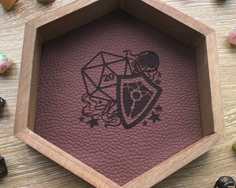 May include: A wooden hexagonal dice tray with a brown leather interior. The tray has an engraved design of a 20-sided die, a shield, and stars. There are several colorful dice scattered around the tray.