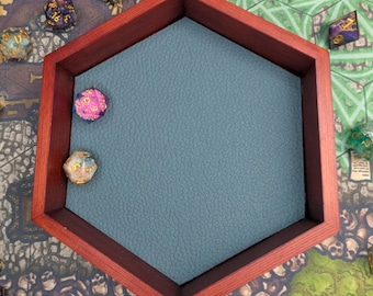 May include: A hexagonal wooden dice tray with a red frame and a teal interior. Several multi-sided dice in various colors are scattered around the tray on a game board with a skull and bone design.