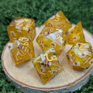 May include: A set of yellow resin dice featuring bees and floral patterns. The dice have gold numerals and are presented on a wooden slice against a green backdrop. The dice are various shapes, including a d20, d12, d10, d8, and d6. The dice are for tabletop games.