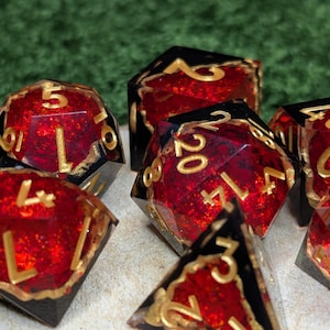 May include: A collection of polyhedral dice with a red, glittery interior and black edges. The numbers are in gold. These dice are designed for tabletop role-playing games. The dice are made of resin.