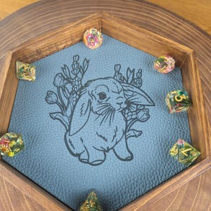 Könnte beinhalten: Ein sechseckiger Würfeltablett aus Holz mit einem blauen Filz-Boden. Der Filz zeigt eine Schwarzweiß-Illustration eines Kaninchens mit Blumen. Das Tablett ist mit bunten Würfeln gefüllt.