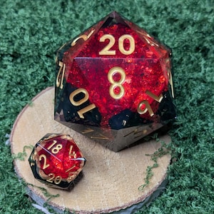 May include: Two polyhedral dice, a D20 and a smaller D6, sit on a wooden slice against a green background. The D20 is large, with gold numerals against a red and black interior. The smaller die has a similar design.