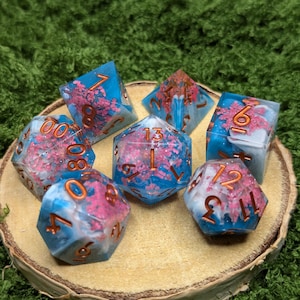 May include: A set of seven polyhedral dice with a blue and white marbled effect, accented with pink floral inclusions and copper-colored numbering. The dice are displayed on a wooden slice against a green background, perfect for tabletop gaming.