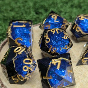 May include: A set of seven polyhedral dice for role-playing games. The dice are black with gold numerals and a blue, glittery interior. The edges are outlined in gold. The dice are displayed on a wooden surface.