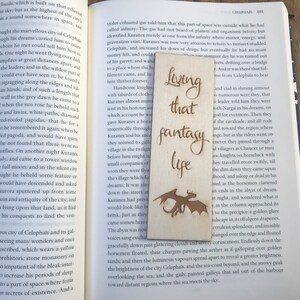 May include: Wooden bookmark with a laser-engraved design of a dragon and the text "Living that fantasy life". The bookmark is light brown with a natural wood grain.