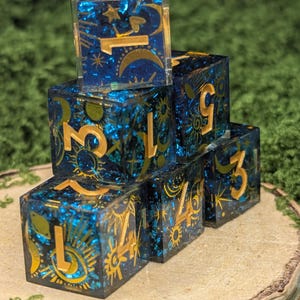 May include: A stack of six translucent blue resin dice with gold-colored numbers and celestial motifs including suns, moons, and stars. The dice have a glittery interior and are arranged in a pyramid formation, displaying various numerical values.