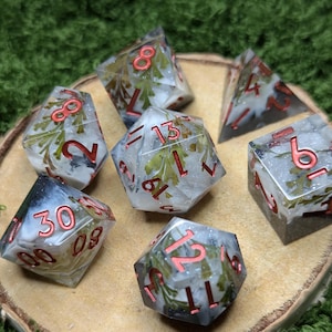 May include: A set of seven polyhedral dice. The dice are clear with white and green inclusions, and red numbering. The dice are displayed on a wooden surface against a green background. The dice are for role-playing games.