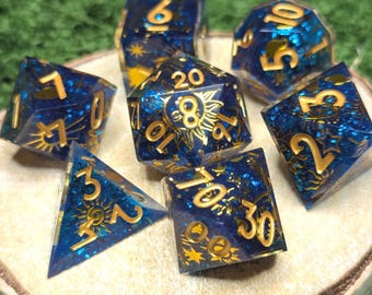 May include: A set of seven polyhedral dice made of translucent dark blue resin with embedded blue glitter. Each die features gold-colored numbers and intricate celestial engravings, including suns, moons, and stars, across all visible faces.