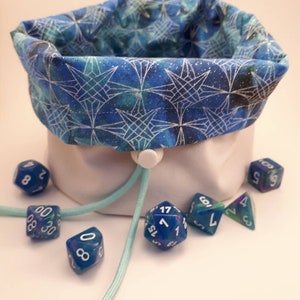 May include: A white fabric dice bag with a blue and white geometric pattern. The bag is open and there are several blue dice with white numbers on them scattered around the bag.