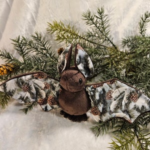 May include: A brown plush bat with a wingspan of approximately 30 cm. The wings are made of a fabric with a woodland print featuring pine trees and pine cones. The bat is sitting on a white fluffy surface.