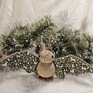 May include: A plush brown bat with green wings featuring a woodland pattern. The wings are decorated with white silhouettes of deer, trees, and other woodland creatures.