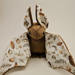 May include: A brown plush bat toy with patterned wings and ears. The wings and ear interiors display a white fabric with a repeating pattern of mushrooms, dragonflies, and ferns. The bat has small black eyes and a soft, furry texture.