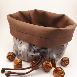 May include: A brown faux leather dice bag with a drawstring closure. The bag is lined with a white fabric featuring a woodland print with deer and trees. The bag is filled with six gold glitter dice.