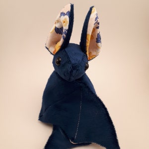 May include: A plush blue bat toy with floral patterned ears. The bat has a stitched seam down the centre of its back.