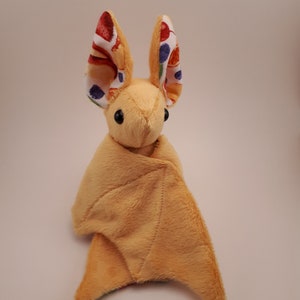 May include: A plush toy bat with a light brown body and large, colourful ears. The ears are white with red, orange, yellow and blue spots.