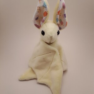 May include: A plush yellow bat toy with a white and colourful ice cream print on its ears. The bat is curled up in a blanket.