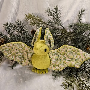 May include: A green plush bat with succulent fabric wings. The bat is sitting on a white fluffy surface with green pine branches.