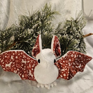 May include: A white plush bat with red and white patterned wings. The wings feature a woodland design with deer, trees, and berries.