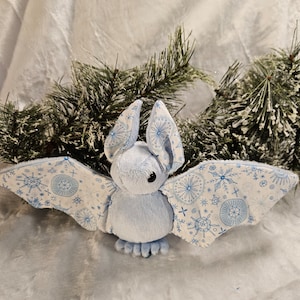 May include: A blue plush bat with snowflake and circle patterns on its wings. The bat is sitting on a white fluffy surface.