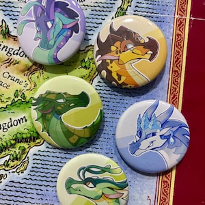 May include: Five circular pinback buttons featuring stylised dragon illustrations. The designs include a purple dragon, a brown and orange dragon, a dark green dragon, a light blue and white dragon, and a light green dragon with long antennae.