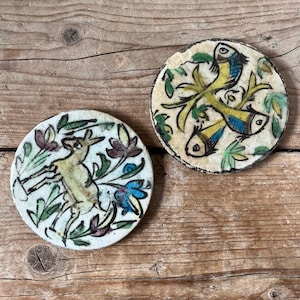 Peut inclure: Deux dessous de verre ronds en céramique avec des motifs peints à la main. L'un représente un cerf entouré de fleurs vertes, violettes et bleues. L'autre présente trois poissons avec des corps jaunes et des détails bleus, sur fond vert et jaune.