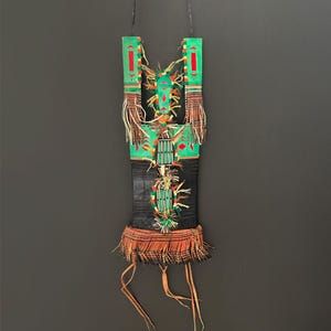May include: A handcrafted, vertical wall hanging with a black base and green top sections. The green sections feature red and yellow geometric designs. The piece is adorned with fringe and tassels in shades of orange, brown, and cream, creating a textured, artistic display.