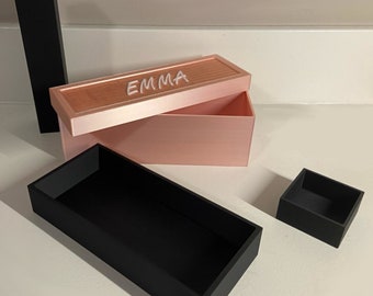 May include: A pink box with a lid that says "EMMA" in white lettering. The box is open and there is a black tray inside. There is also a smaller black tray to the right of the box.