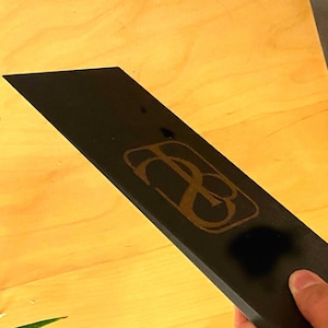 May include: A black, rectangular object with a gold-colored logo. The logo appears to be a stylized monogram or emblem. The object is held by a hand, and the background is a light-colored wooden surface.