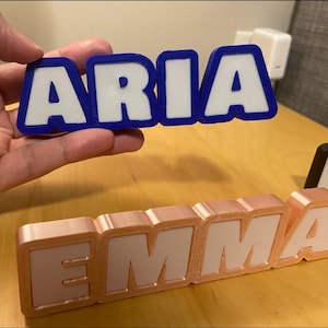 Könnte beinhalten: 3D-gedruckte Namensschilder. Ein Schild schreibt "ARIA" in weißen Buchstaben mit blauem Rand, gehalten von einer Hand. Ein weiteres Schild schreibt "EMMA" in weißen Buchstaben mit roségoldenem Rand. Ein drittes Schild mit schwarzem Rand ist teilweise sichtbar.